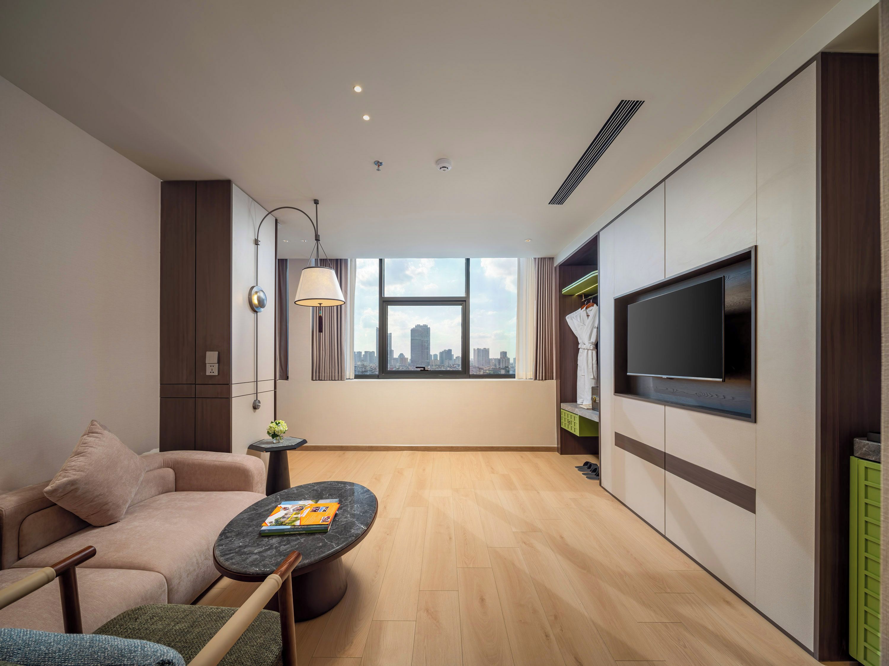 Habitación de hotel moderna con sofá, una mesita redonda con una revista, televisor montado en la pared y ventana con vista a la ciudad.