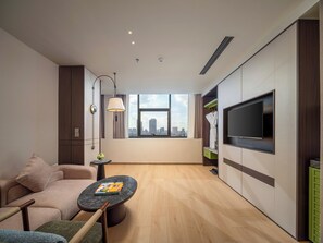 Ett modernt hotellrum med en soffa, ett litet runt bord med ett tidningsnummer, en tv som är monterad på väggen och ett fönster med utsikt över staden.