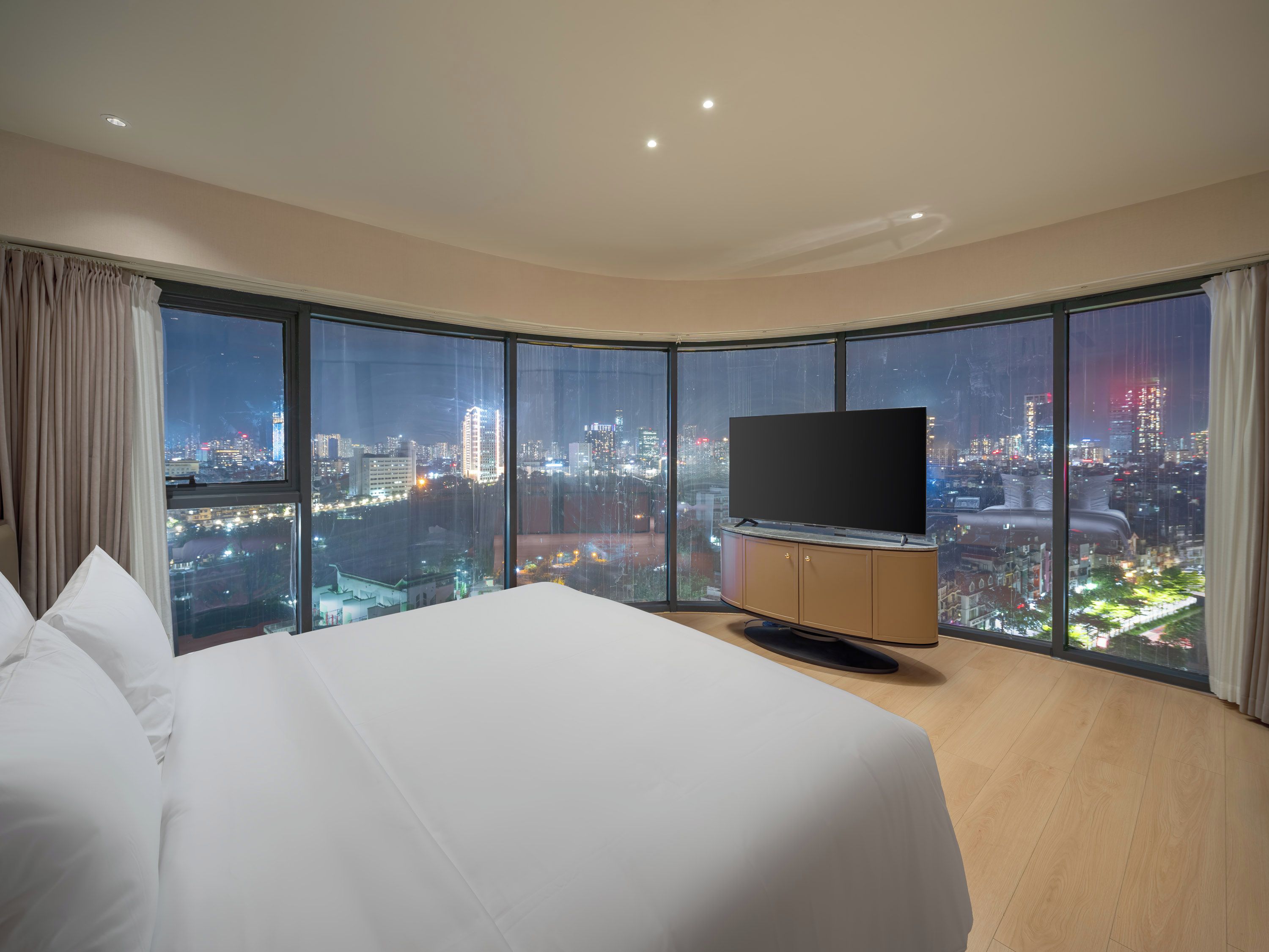 A hotel room with a large bed, a flat-screen TV, and a city view through large windows.