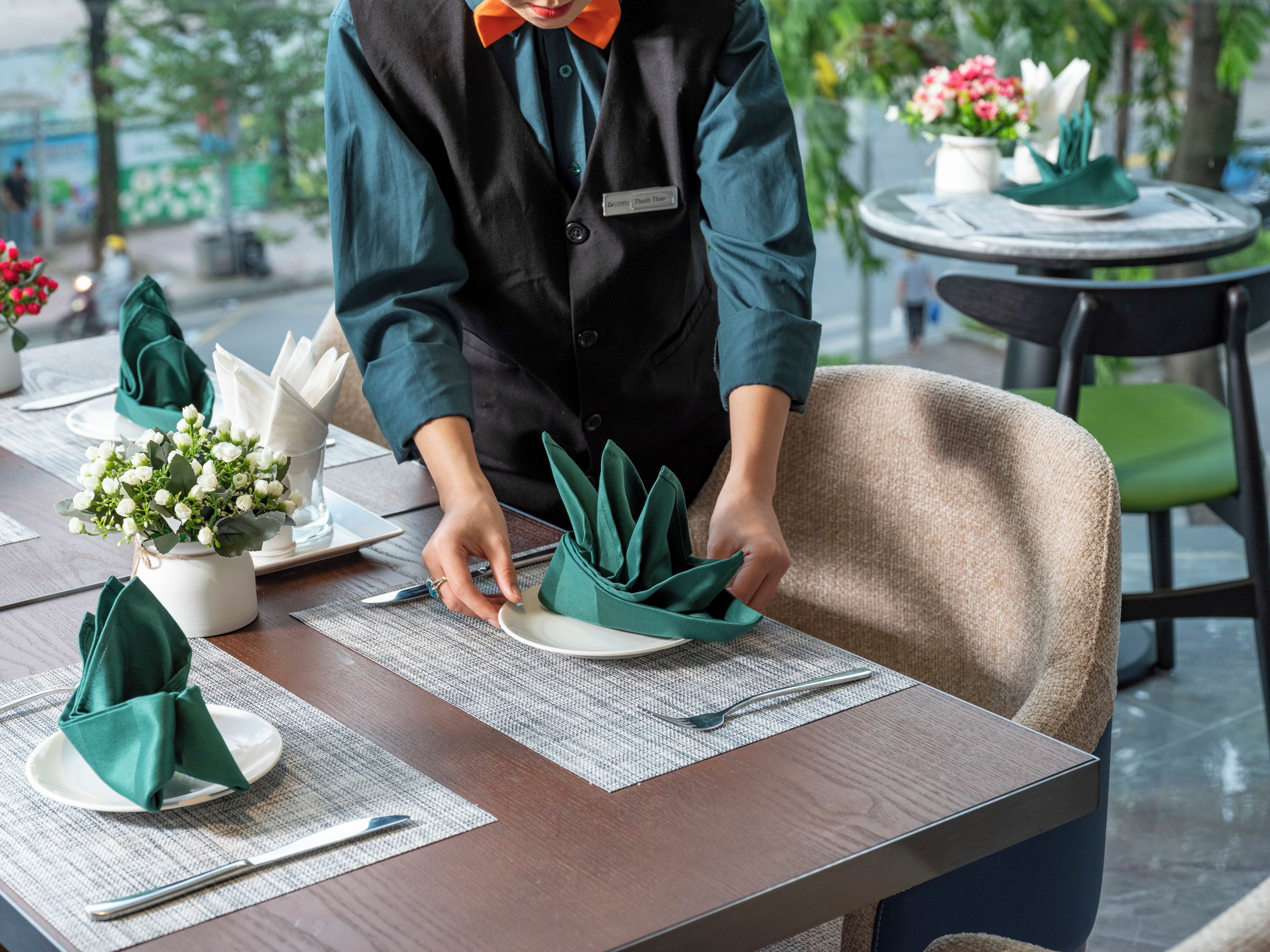 Une personne prépare une table en y disposant des serviettes vertes et des assiettes blanches, un petit vase blanc avec des fleurs et un badge nominatif.