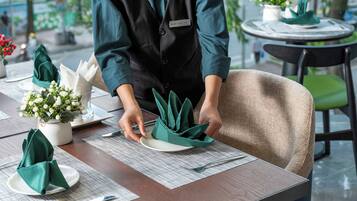 Une personne prépare une table en y disposant des serviettes vertes et des assiettes blanches, un petit vase blanc avec des fleurs et un badge nominatif.