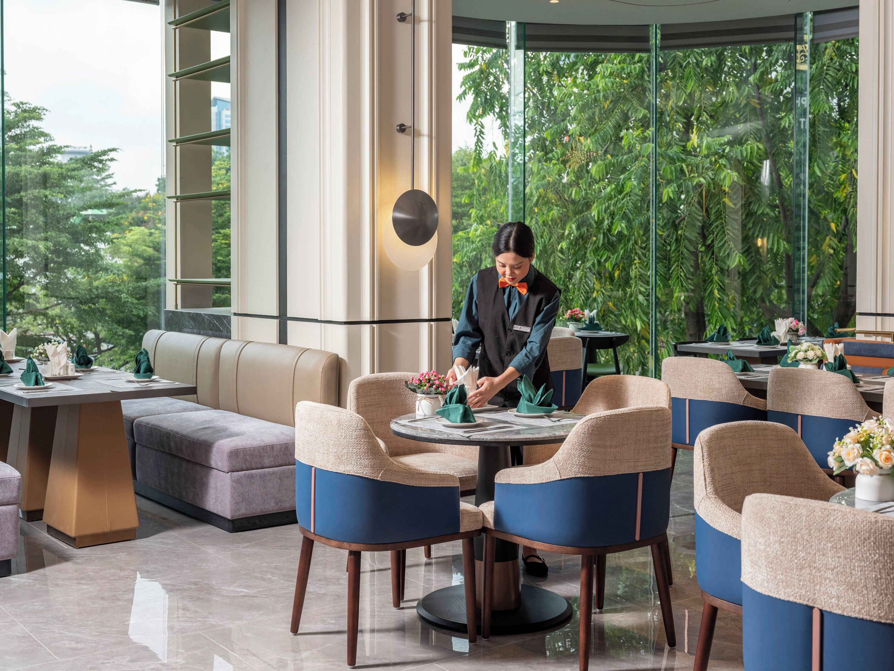 A modern dining area with a waiter serving at a table, surrounded by comfortable seating and large windows offering a view of greenery.