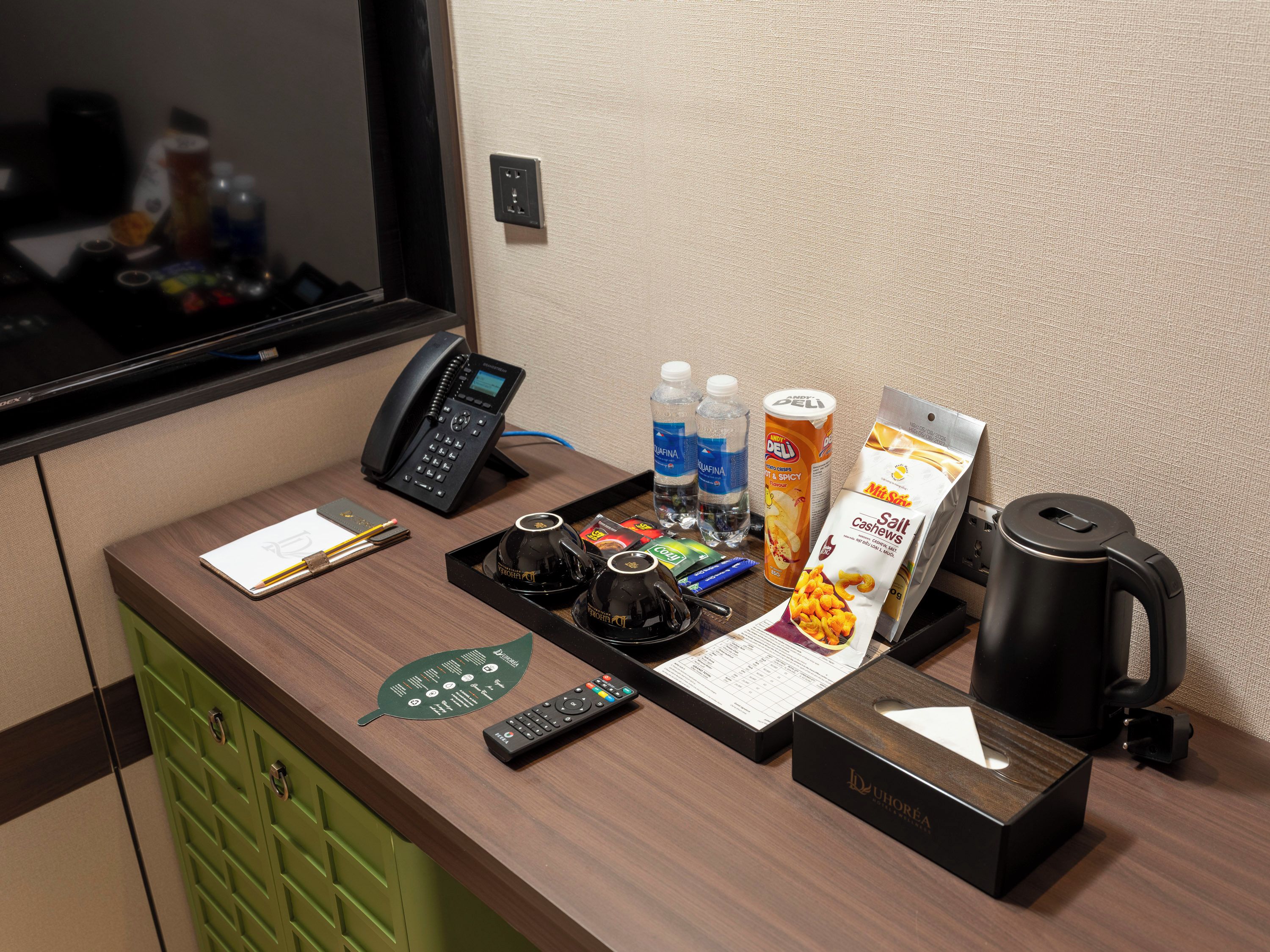A hotel reception desk with a telephone, remote controls, a menu, bottled water, a salt packet, and a coffee maker.
