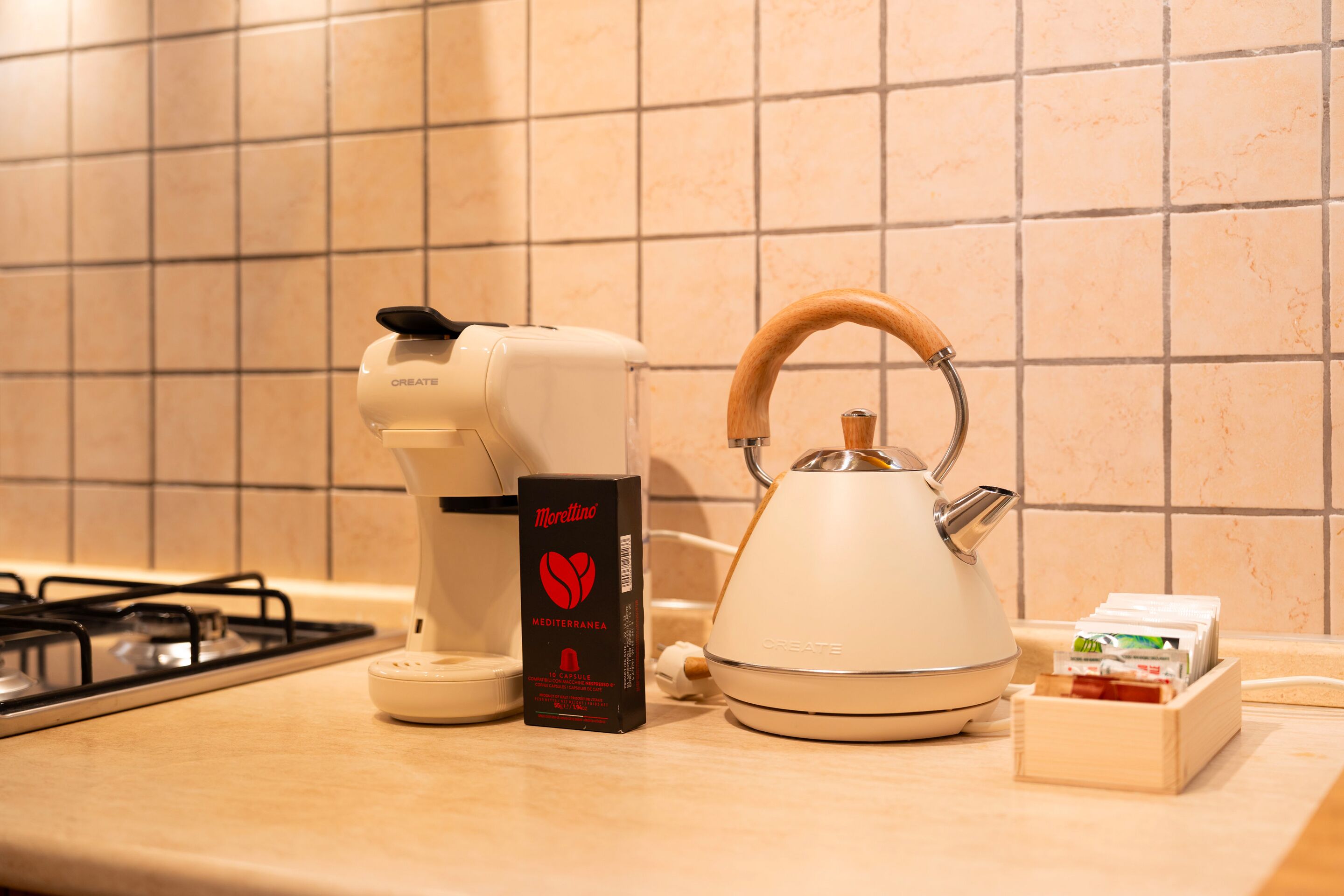 A kitchen with a gas stove, a coffee maker, a kettle, and a box of Morellino coffee.