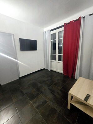 Ein Zimmer mit dunklem Fliesenboden, einer weißen Tür, einem an der Wand befestigten Flachbildfernseher und einem Fenster mit roten Vorhängen.
