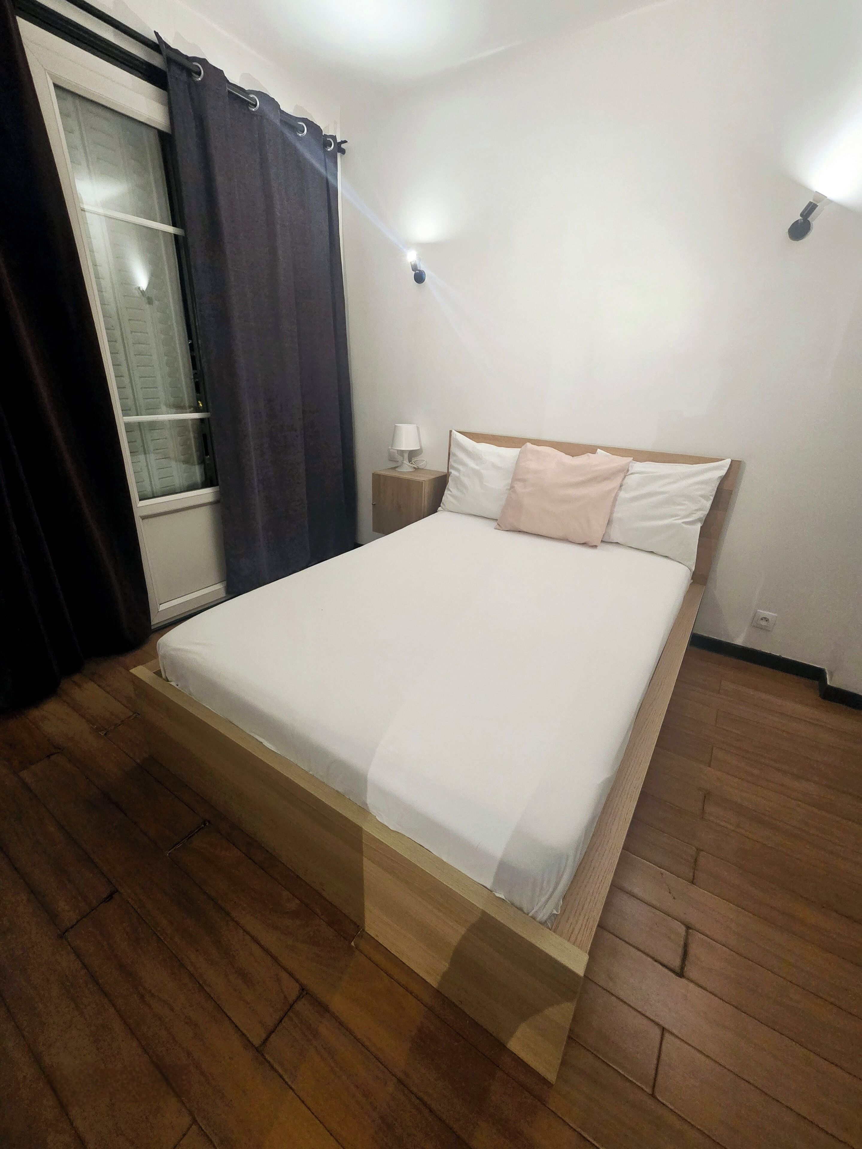 Ein ordentlich bezogenes Bett mit Holzbettgestell, ein Nachttisch mit Lampe und ein Fenster mit Vorhängen.