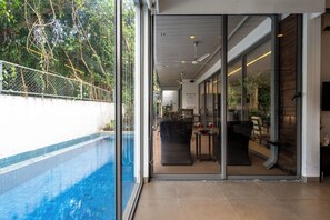 A modern indoor pool area with a glass wall, a ceiling fan, and a seating area with a table and chairs. - Staymaster Belamia•3BR•Private Pool (Parra)