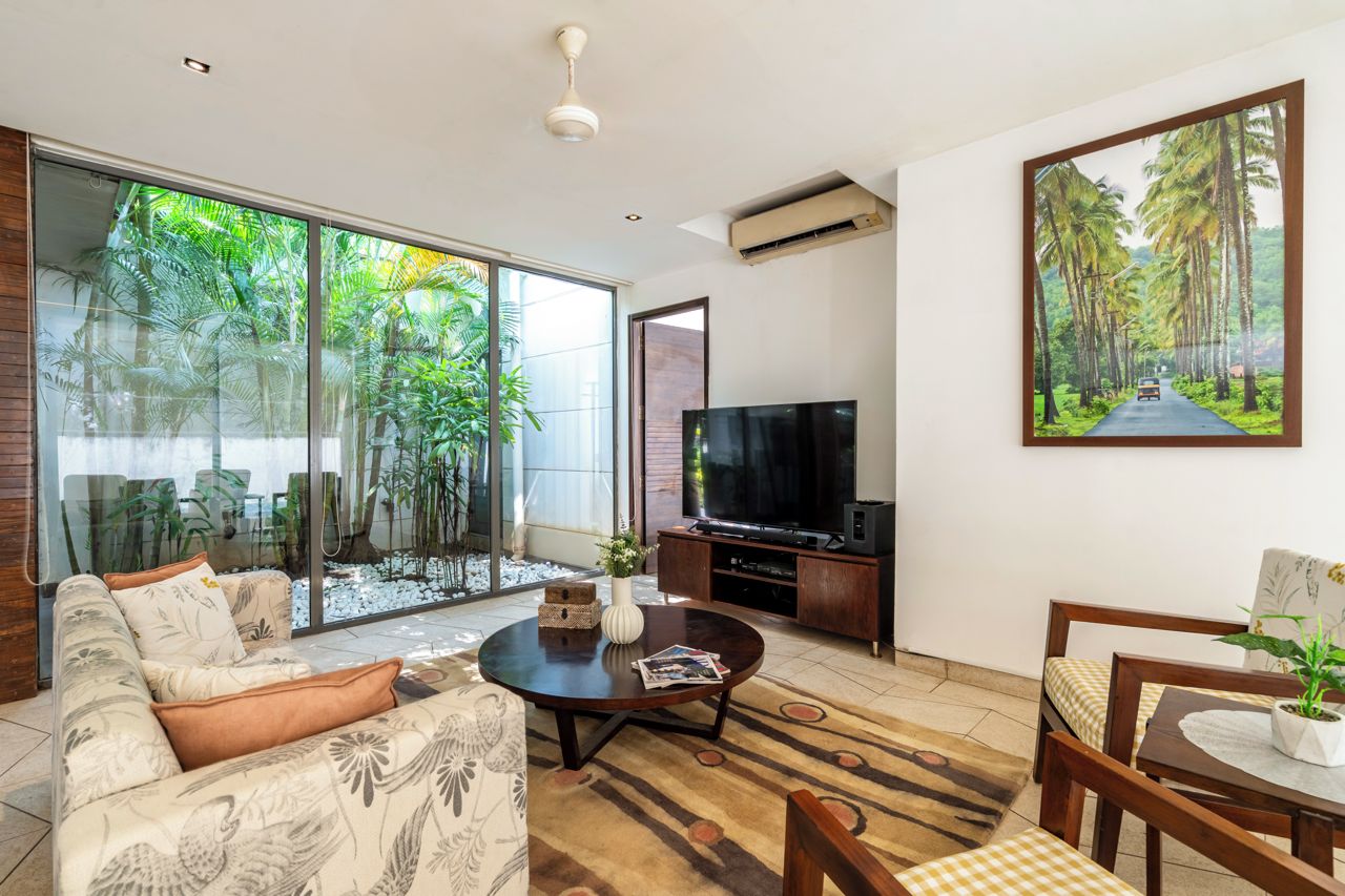 Een moderne woonkamer met een bank, salontafel en televisie, grote glazen schuifdeuren die leiden naar een balkon met planten, en een schilderij van een tropische weg.