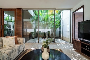 A living room with a sofa, a coffee table with a vase, a flat-screen TV, and a sliding glass door leading to a garden. - Staymaster Belamia•3BR•Private Pool (Parra)