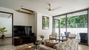 A modern living room with a flat-screen TV, comfortable seating, a potted plant, and a large window offering a view of greenery. - Staymaster Belamia•3BR•Private Pool (Parra)