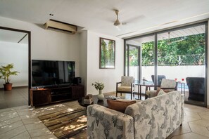 A modern living room with a flat-screen TV, comfortable seating, a potted plant, and a large window offering a view of greenery. - Staymaster Belamia•3BR•Private Pool (Parra)