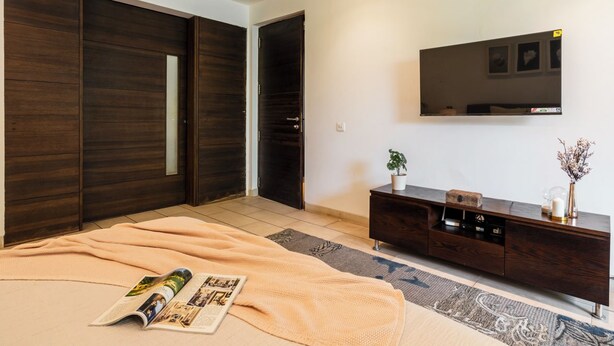 A bedroom with a large bed, a dark wooden wardrobe, a TV mounted on the wall, and a small side table with decorative items. - Staymaster Belamia•3BR•Private Pool (Parra)