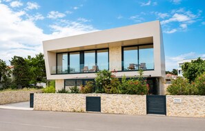 A modern two-story building with large windows, a balcony with seating, and a stone wall in front. - Gorgeous home in Novalja (Novalja)
