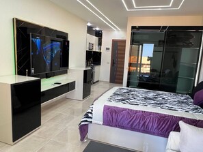 A modern bedroom with a large bed, a TV mounted on the wall, a built-in kitchen area, and a sliding glass door closet. - Modern Studio Apartment With Stunning Red Sea Views (Hurghada)