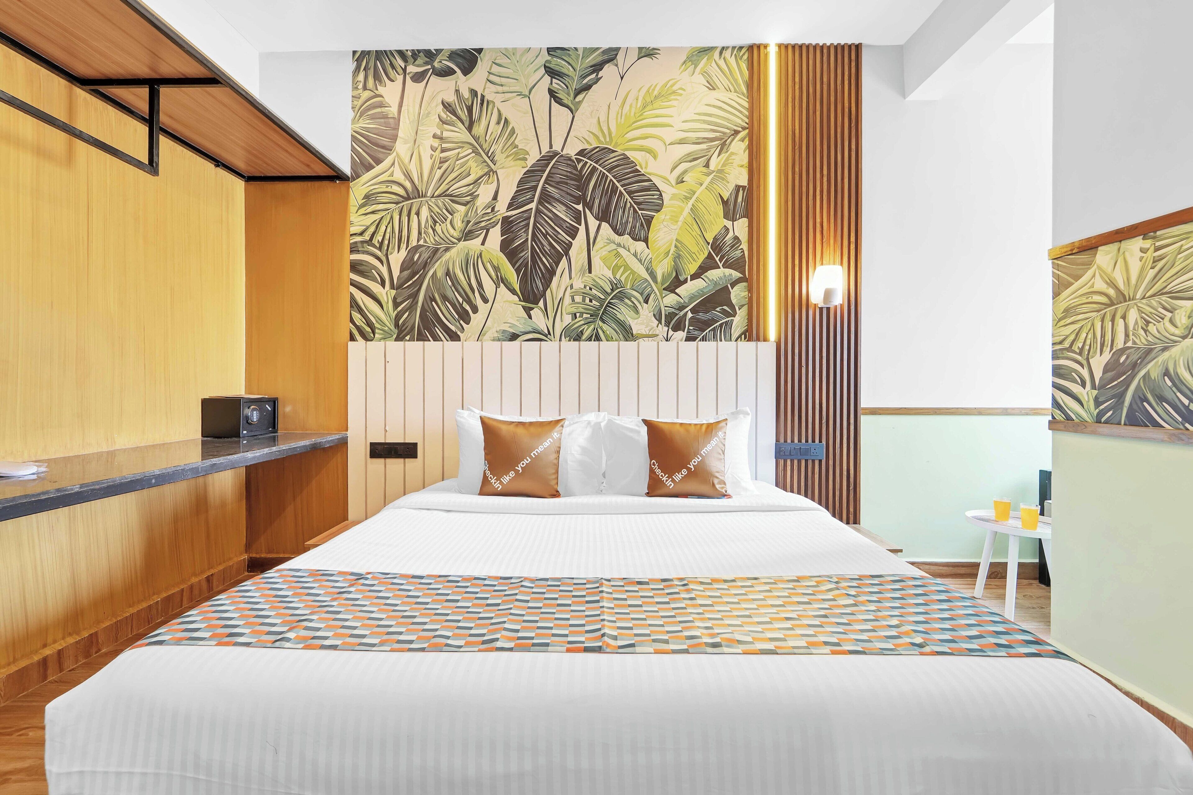 Quarto de hotel com uma cama, uma escrivaninha de madeira, uma parede com padrão de folhagem tropical e uma pequena mesa com duas chávenas.