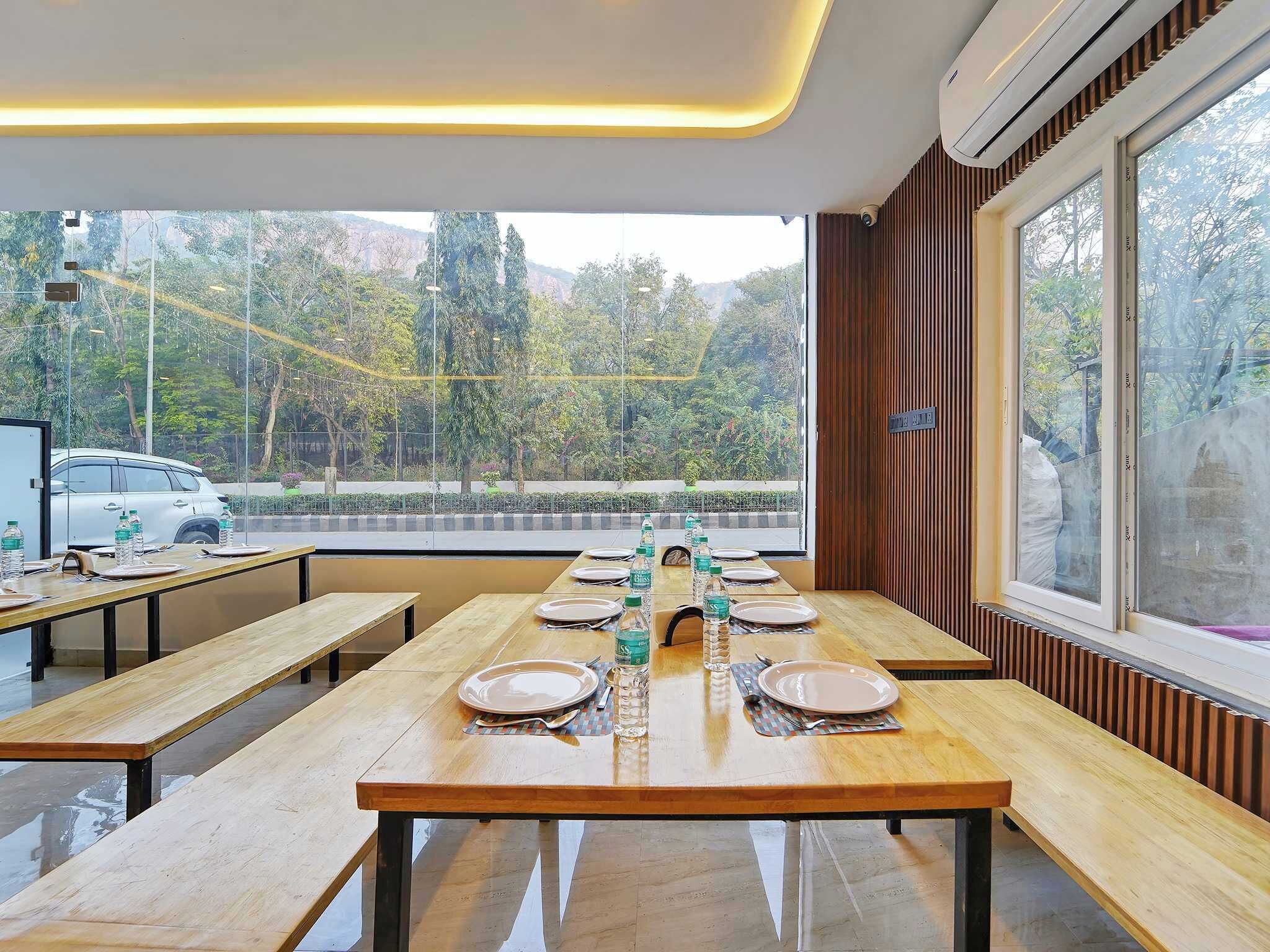 Un comedor con mesas y bancos de madera, vistas a árboles y una carretera fuera, y un equipo de aire acondicionado en la pared.