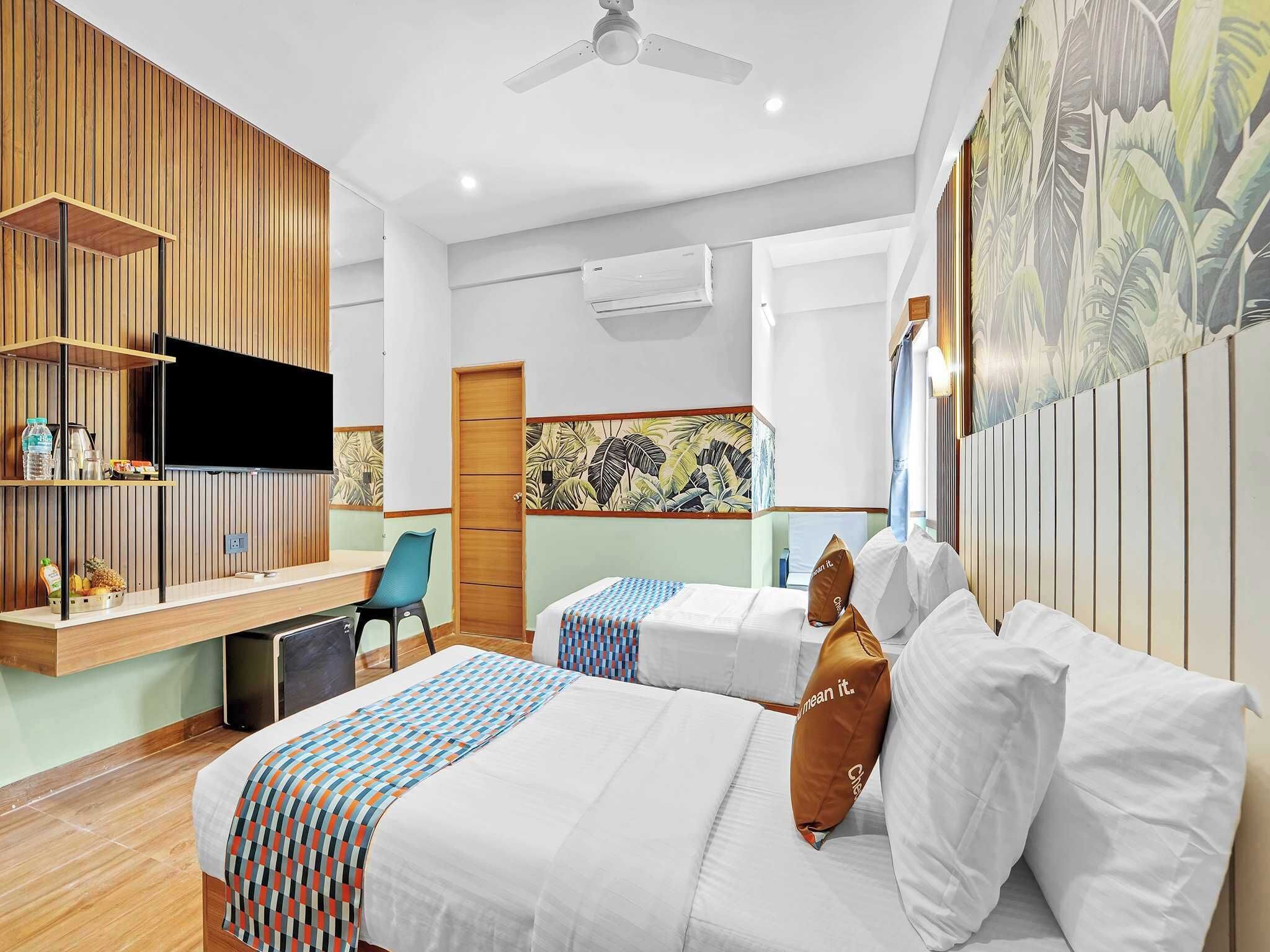 A hotel room with a bed, a desk with a flat-screen TV, a chair, and a wall with a tropical leaf pattern.