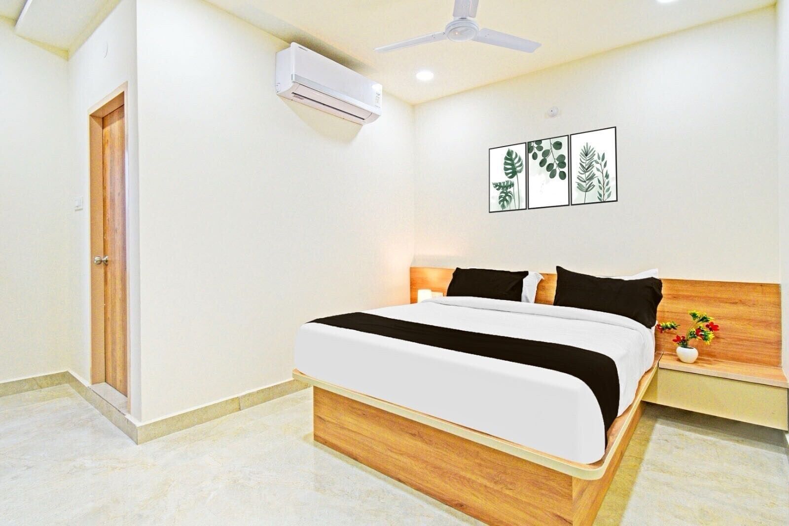 A hotel room with a bed, a nightstand, a wall-mounted air conditioner, a ceiling fan, and a framed botanical artwork.