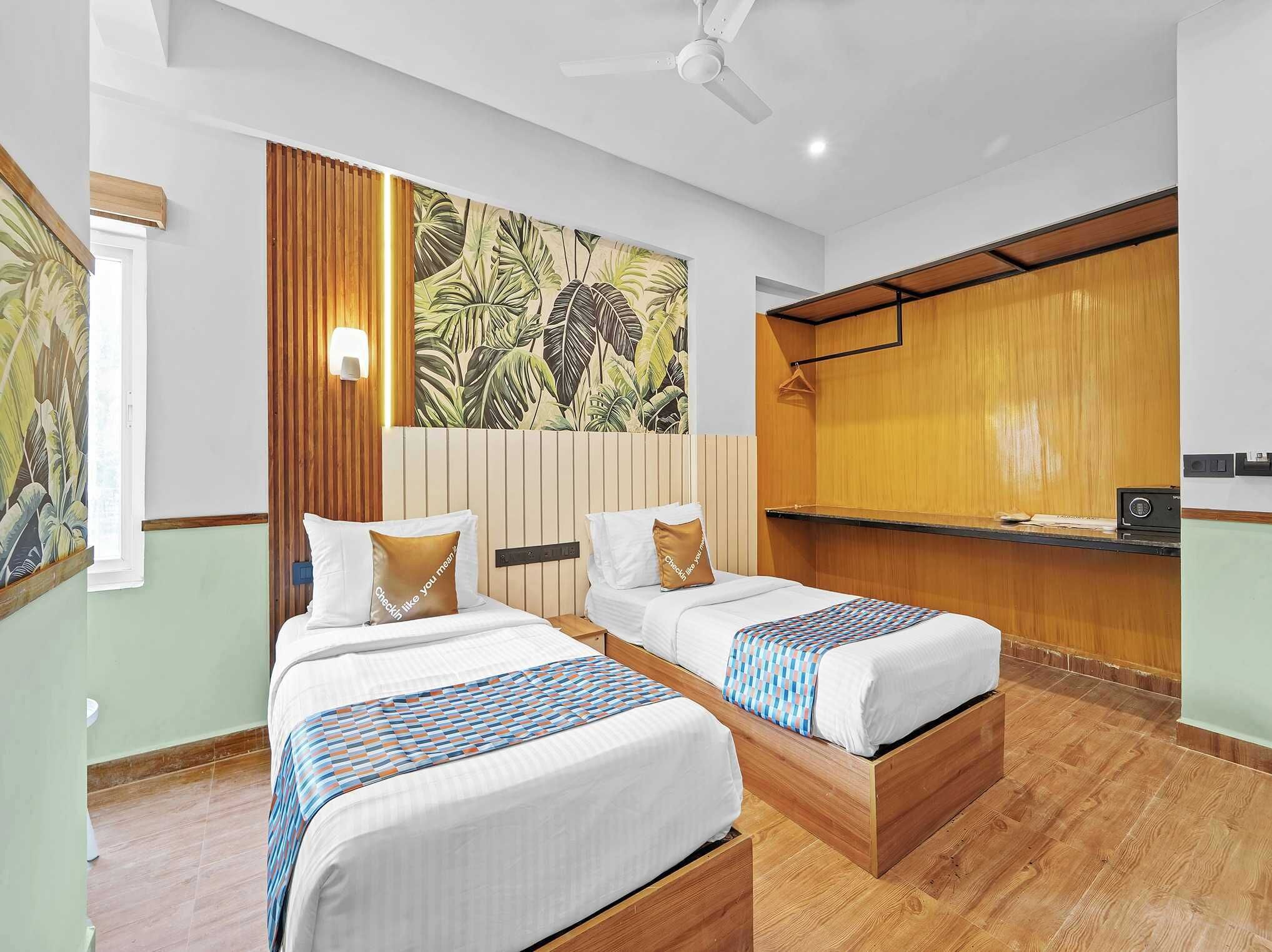 Habitación de hotel con dos camas, cabecero de madera, pared con estampado de hojas tropicales, ventilador de techo y una pequeña kitchenette.