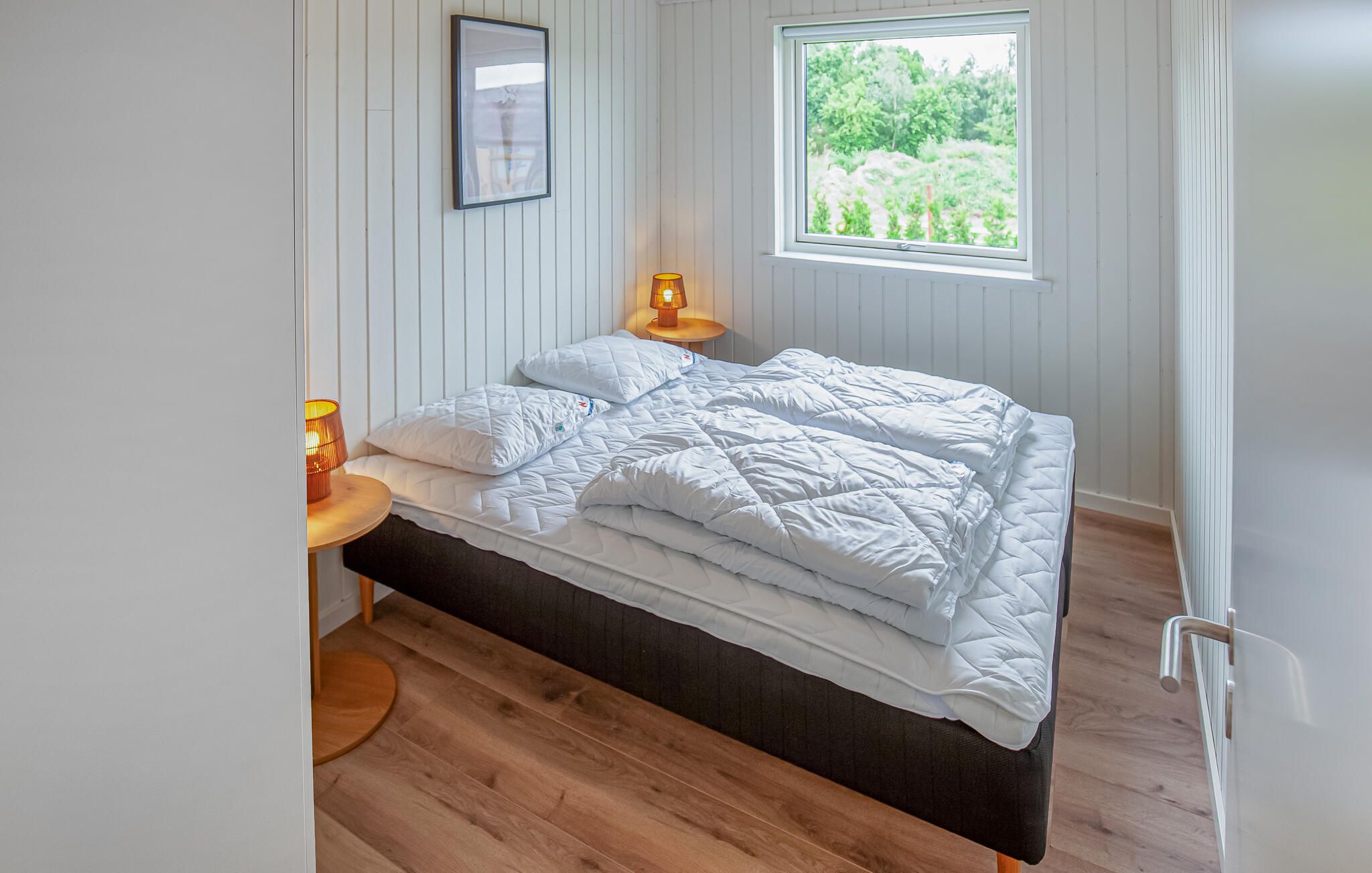 A bedroom with a bed, two bedside lamps, a window with a view of greenery, and a framed picture on the wall.