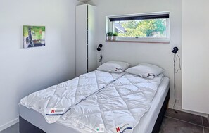 Un lit simple avec une couette blanche, une petite fenêtre donnant sur la verdure et une lampe fixée au mur.