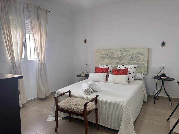 Quarto bem arrumado com uma cama, uma cadeira, uma mesinha de cabeceira e uma pequena mesa com abajur.