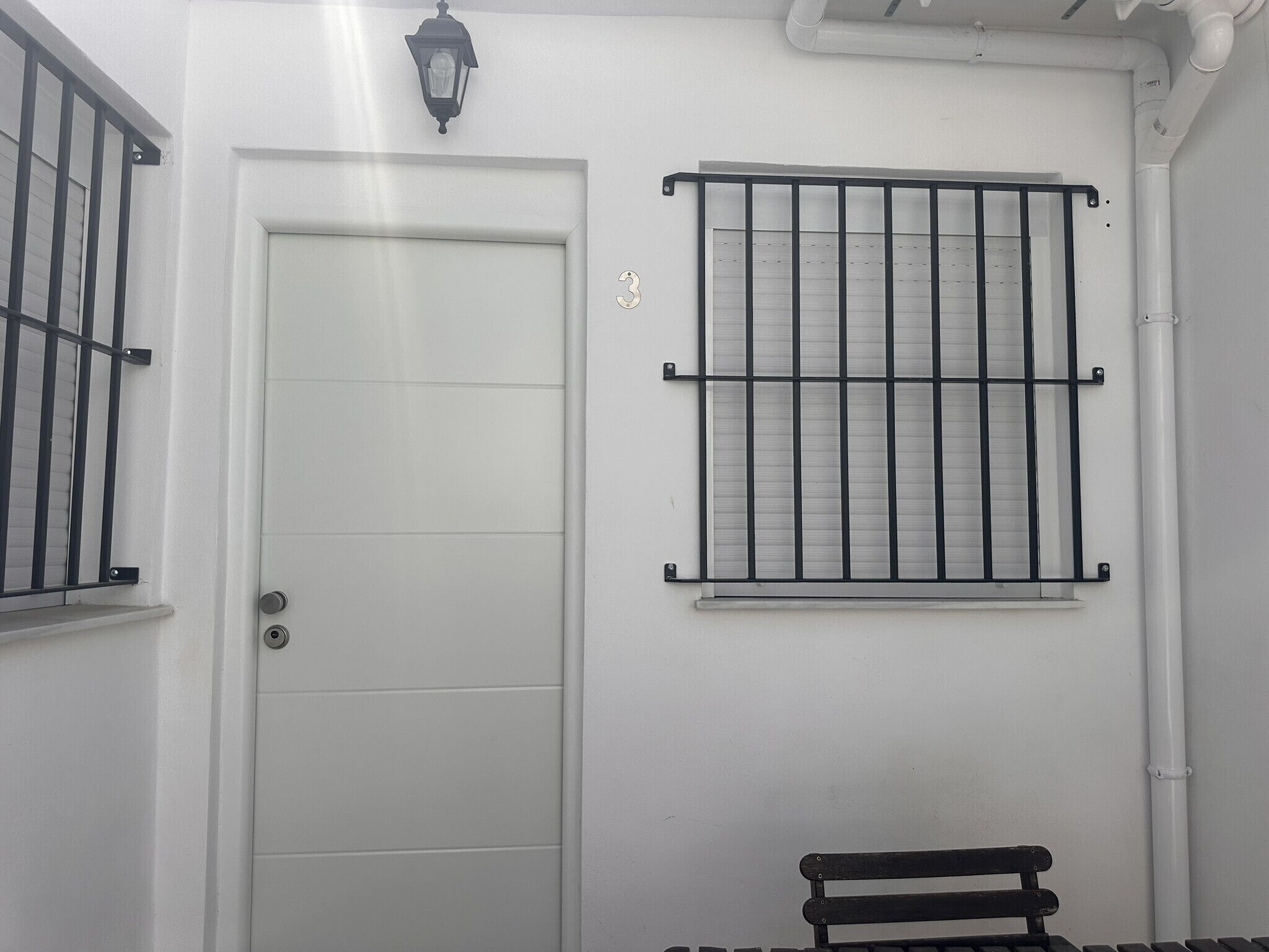 A white door with a metal gate, a window with metal bars, and a bench.
