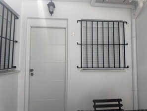 Uma porta branca com grade de metal, uma janela com barras de metal e um banco.