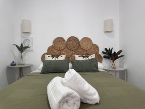 Ein ordentlich gemachtes Bett mit einer grünen Bettdecke, zwei grünen Kissen und einem weißen Handtuch. An der Wand hinter dem Bett befinden sich zwei runde Wanddekorationen aus gewebtem Material.