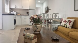 Ett modernt kök med vita skåp, en matplats med ett träbord och stolar samt ett vardagsrum med en soffa och dekorativa kuddar.