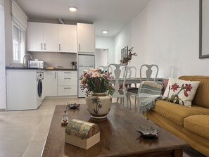 Eine moderne Küche mit weißen Schränken, ein Essbereich mit Holztisch und -stühlen sowie ein Wohnzimmer mit Sofa und Zierkissen.