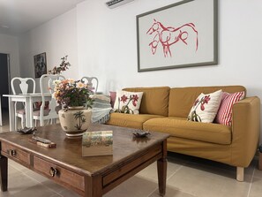 Uma sala de estar com um sofá marrom, cadeiras brancas, uma mesa de centro de madeira e um vaso com flores.