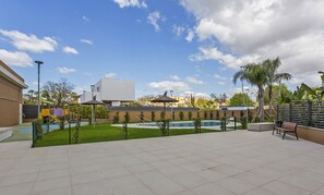 Un espace piscine sur le toit, avec une piscine entourée d’une clôture, une aire de jeux pour enfants et un coin salon équipé d’un banc.
