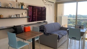 A modern living room with a sofa, a dining table, chairs, a TV, and a balcony with a view. - Large Studio in Canne Marina (Mandelieu-la-Napoule)