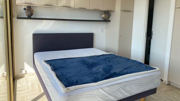 A bedroom with a bed, a headboard, a shelf with decorative items, and a door. - Large Studio in Canne Marina (Mandelieu-la-Napoule)