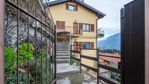 Ein Wohngebäude mit einer Treppe, die zu einem Balkon führt, umgeben von viel Grün und einer Bergkulisse.