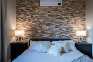 A neatly made bed with white linens and pillows, a stone-patterned wall, and two bedside lamps on nightstands. - Limonta Breathless View by Wonderful Italy (Limonta)