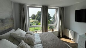 Ein Schlafzimmer mit einem großen Bett, zwei mit Vorhängen versehenen Fenstern, einem Heizkörper und einem an der Wand befestigten Fernseher.