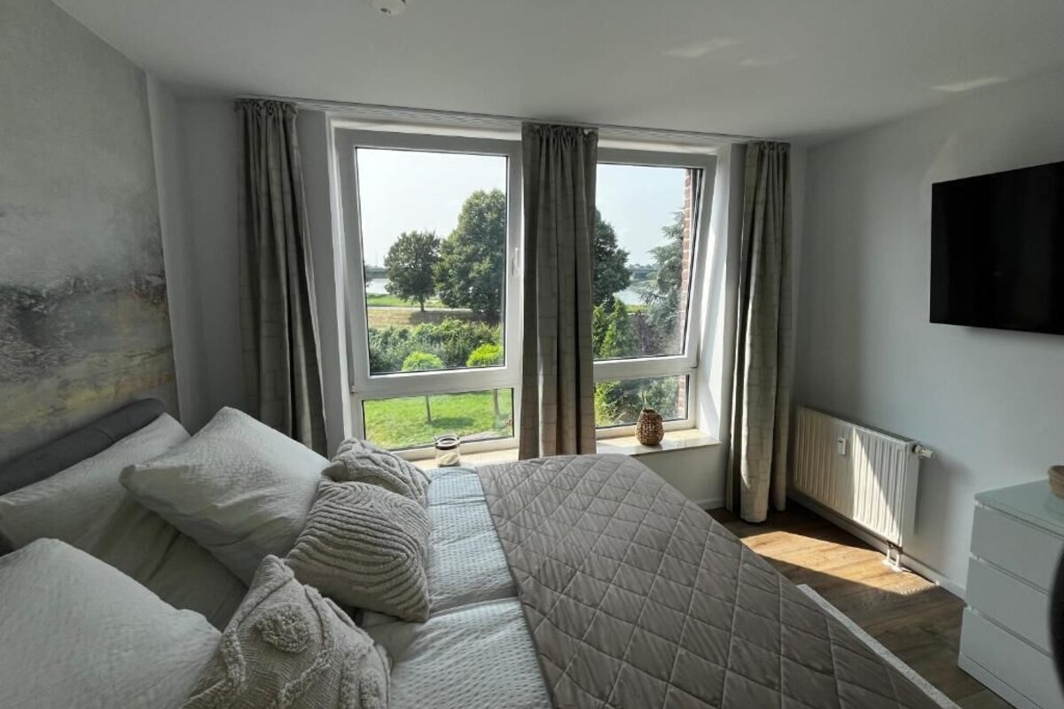 Ein Schlafzimmer mit einem großen Bett, zwei mit Vorhängen versehenen Fenstern, einem Heizkörper und einem an der Wand befestigten Fernseher.