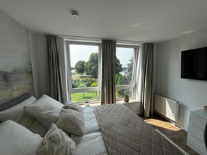 Ein Schlafzimmer mit einem großen Bett, zwei mit Vorhängen versehenen Fenstern, einem Heizkörper und einem an der Wand befestigten Fernseher.