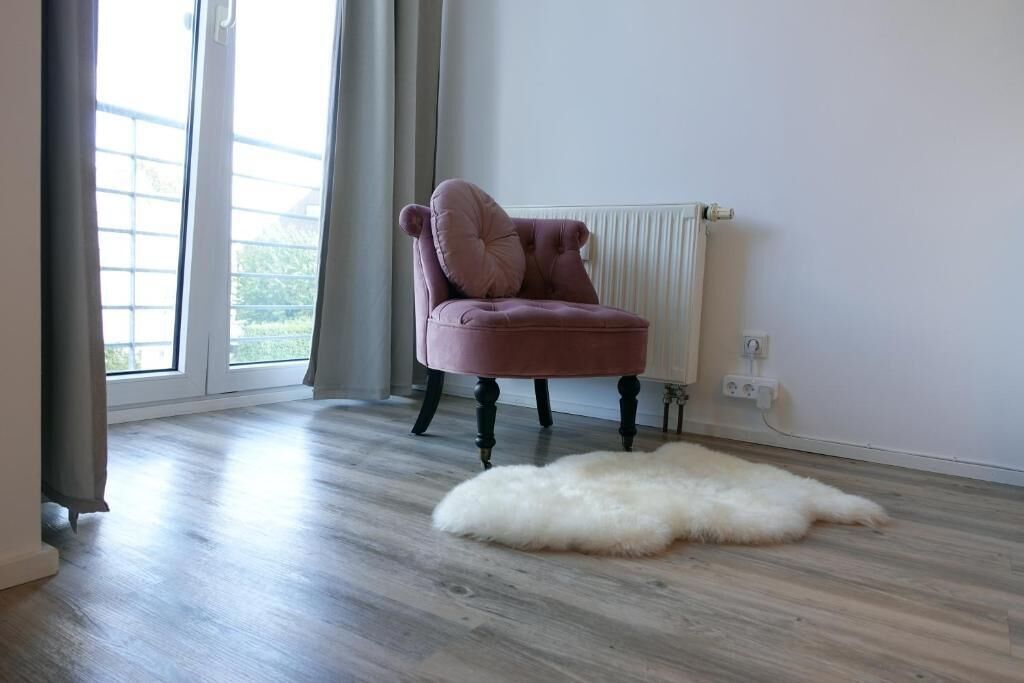 Ein Zimmer mit großem Fenster, einem bequemen Sessel, einem weißen, flauschigen Teppich und einem Heizkörper.