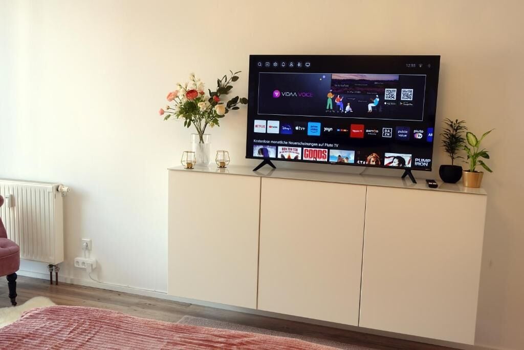 Ein Flachbildfernseher an einem weißen Schrank, daneben eine Blumenvase und eine kleine Pflanze.