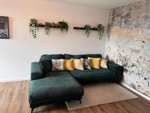 A green sectional sofa with patterned cushions, a wooden floor, a textured wall, and a hanging plant shelf.