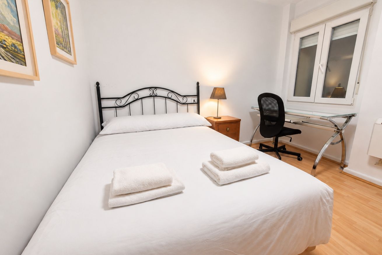 Un lit bien fait, avec du linge de lit blanc et deux serviettes pliées, une lampe de chevet, un fauteuil de bureau noir et une fenêtre à vitres dépolies.