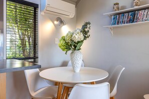 Un coin repas avec une table ronde, des chaises blanches, un vase avec des fleurs, une fenêtre avec des stores, un climatiseur et une bibliothèque remplie de livres.