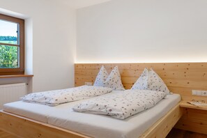 A double bed with floral bedding, a wooden headboard, a bedside table with glasses, and a window with a view of greenery. - Gatterpunerhof "Völs" Farmhouse with Mountain View, Shared Terrace and Wi-Fi (Völs am Schlern)