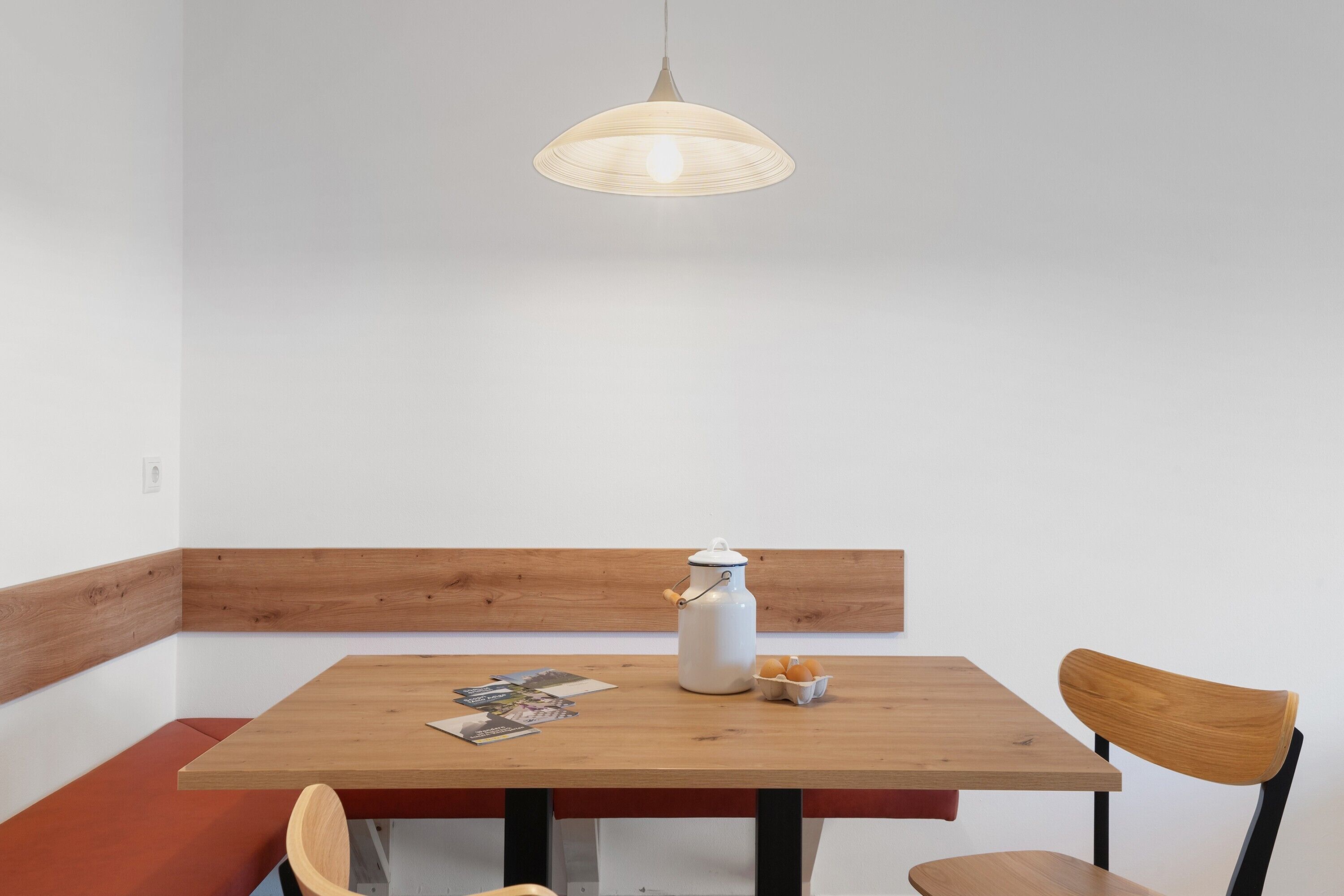 Une table en bois avec un pichet blanc et des œufs, une chaise en bois et un luminaire suspendu.