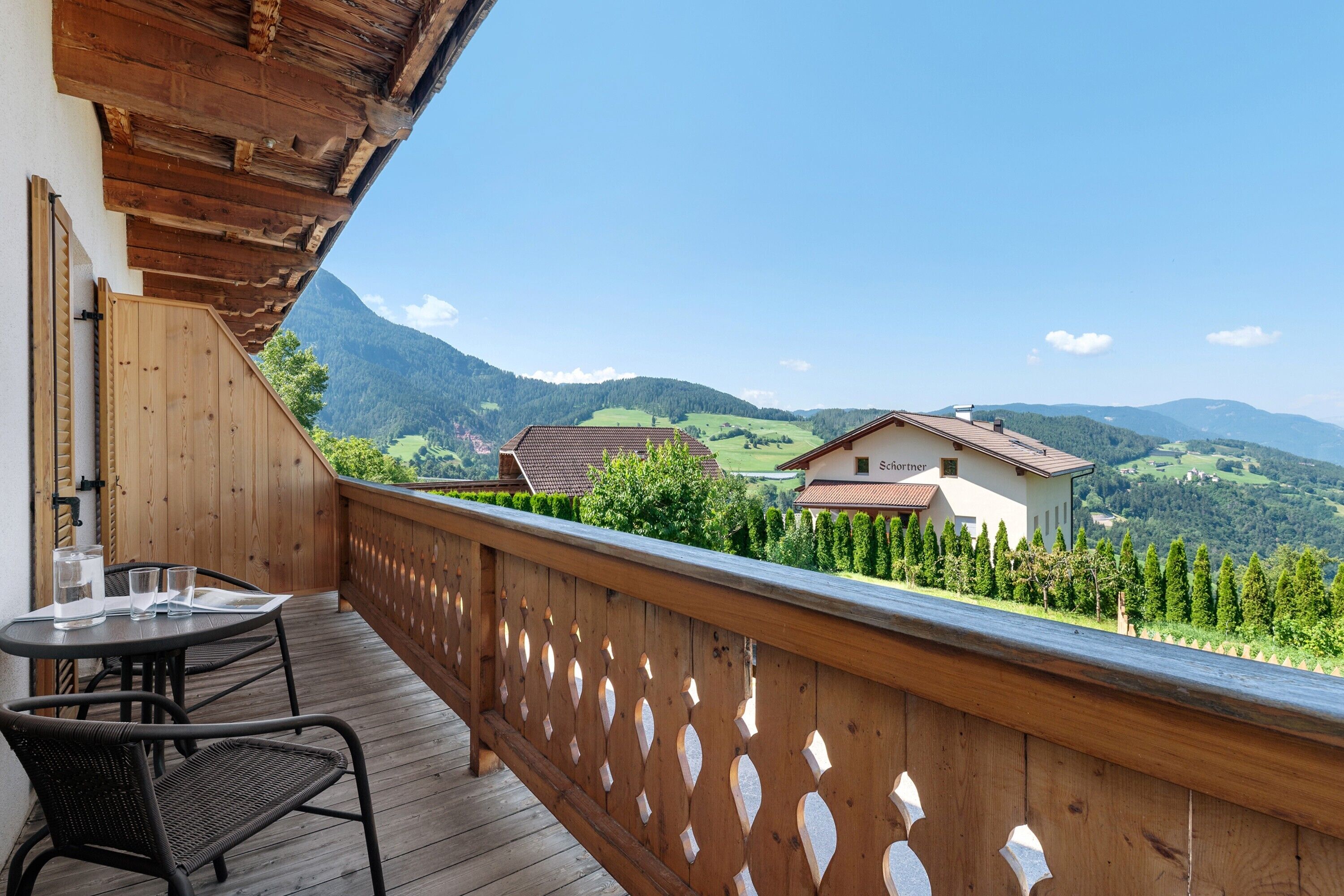 Een houten balkon met een tafel en stoelen, met uitzicht op een bergachtig landschap en een gebouw met de tekst 'Schorner'.