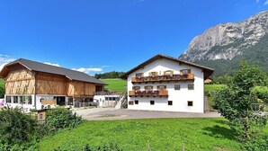 Un edificio de dos pisos con un balcón y una estructura similar a un granero al lado, situado en un paisaje rural con montañas al fondo.