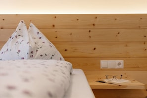 Ein hölzerner Kopfseitenverkleidung mit Steckdose, ein Bett mit blumigem Bettzeug und ein Nachttisch mit Gläsern und einem Buch.